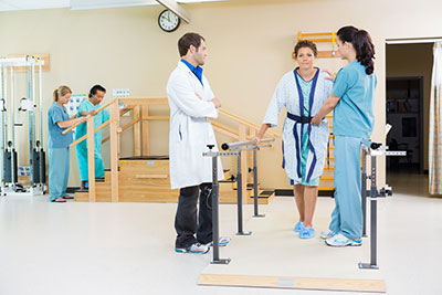 A nurse is helping a patient with physical therapy while a doctor is watching the patient try to walk. While in the background there is another nurse helping a patient walk up the stairs